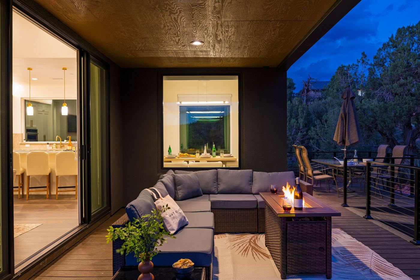 The covered lounge at dusk with the fire table lit and the kitchen glowing through the glass doors.